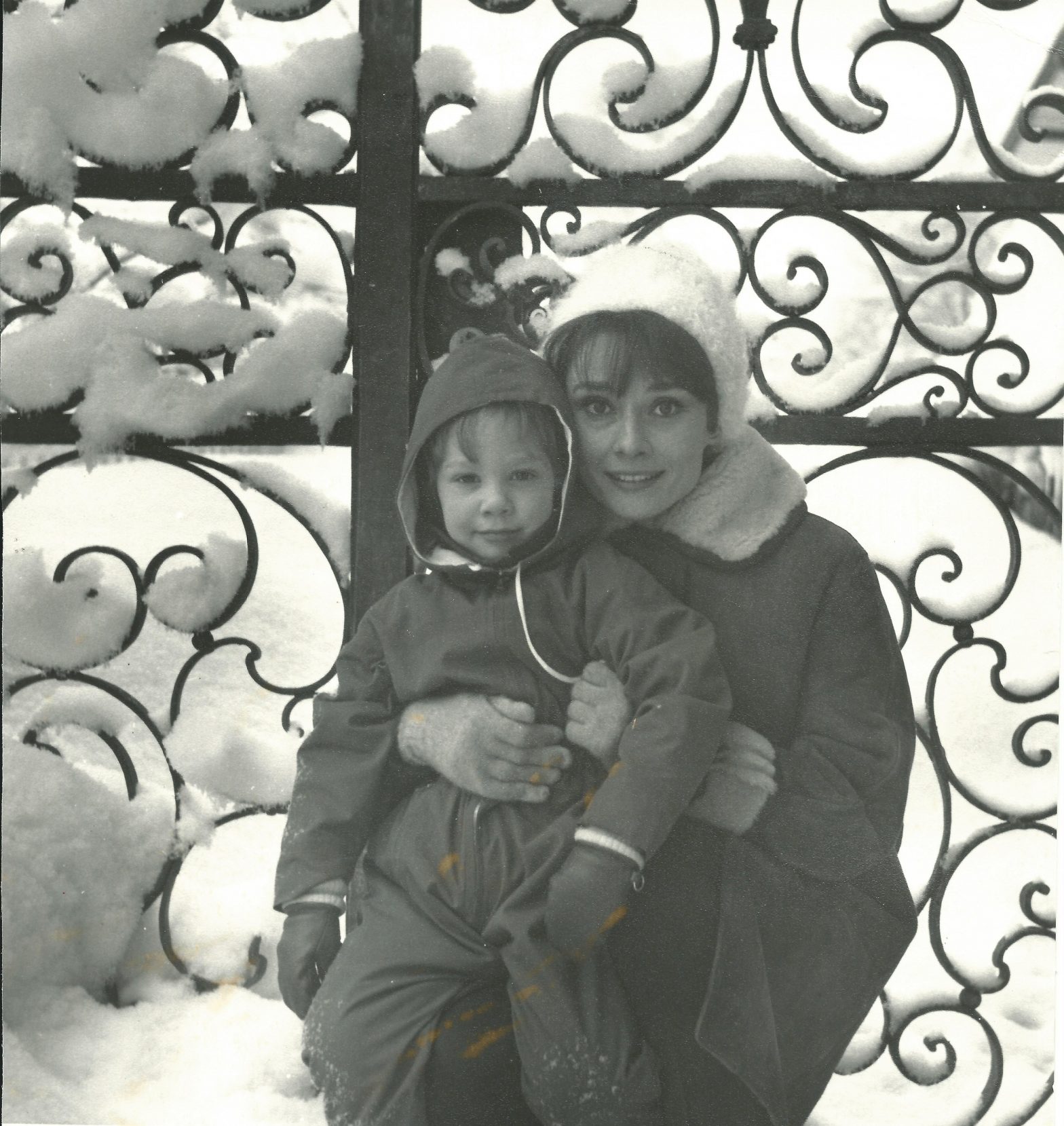 An image of Audrey Hepburn with Sean Hepburn Ferrer on her lap sitting in the snow against a fence in black and white.