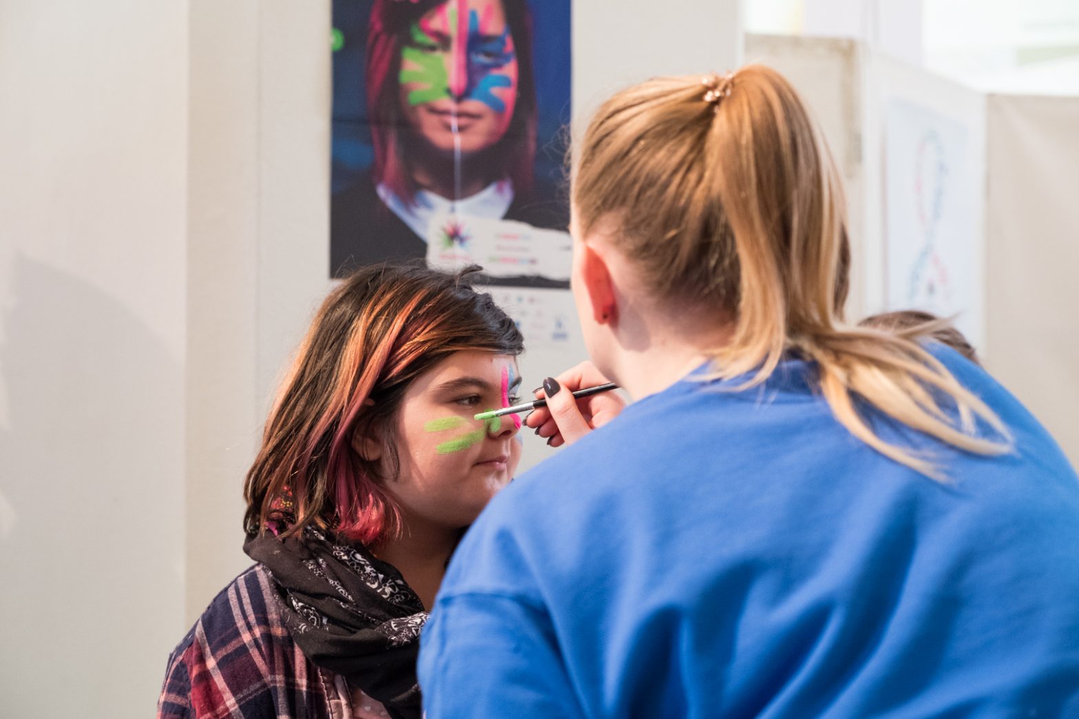 An image of a girl's face being painted with rare disease day stripes in green, pink and blue.